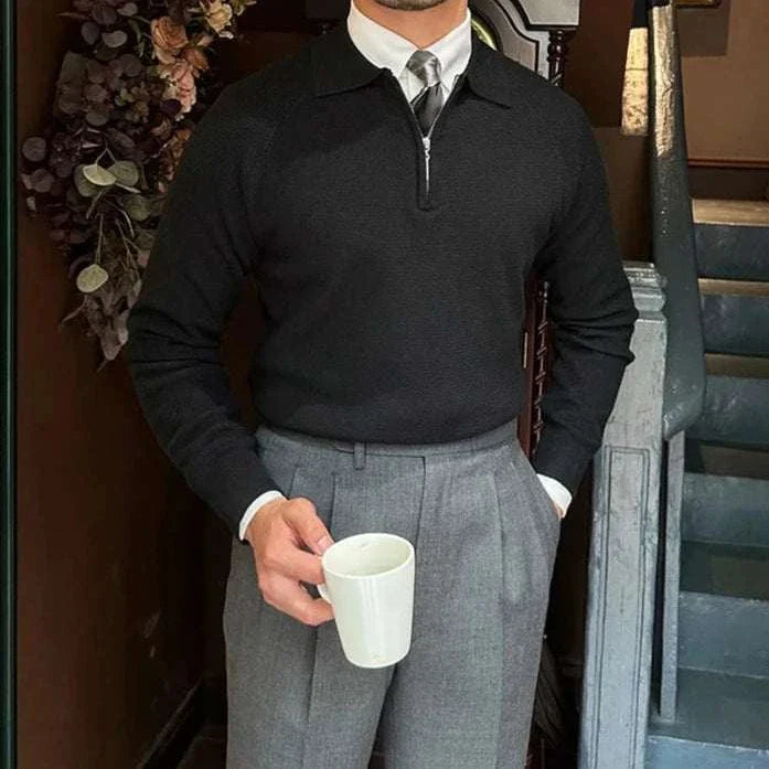 Man wearing an Oxford zipper sweater in black with a solid pattern, styled with gray trousers, holding a coffee mug indoors.