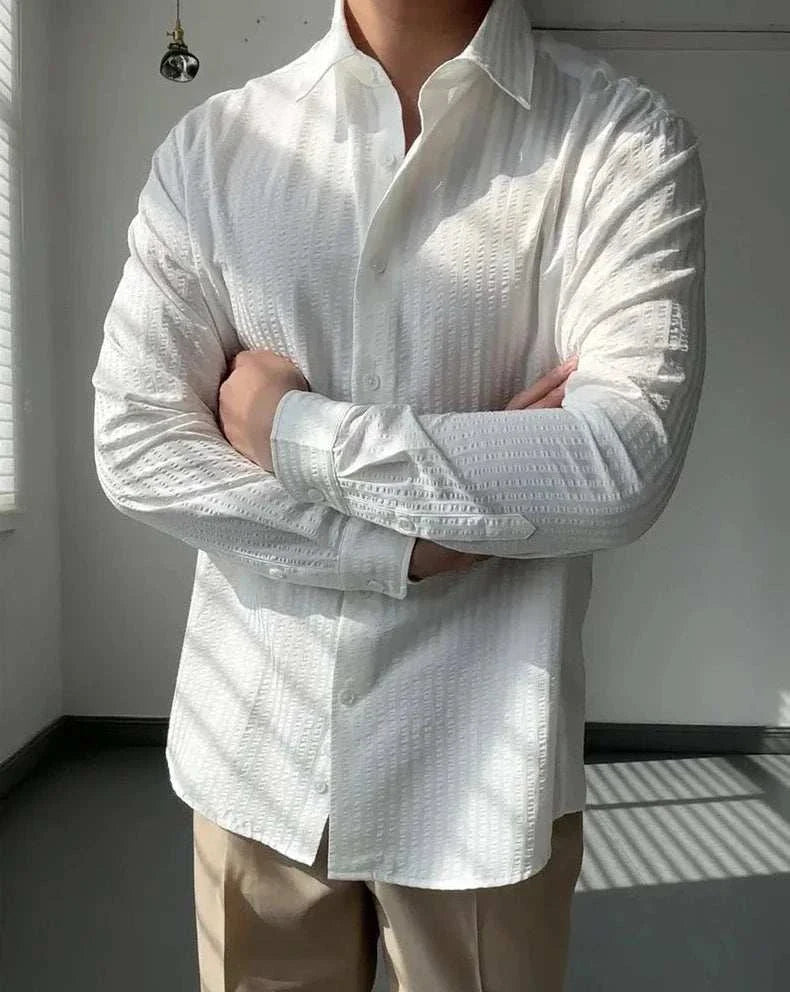 Men's white striped shirt with classic collar, Antonios British casual style, paired with beige pants in a sunlit room.