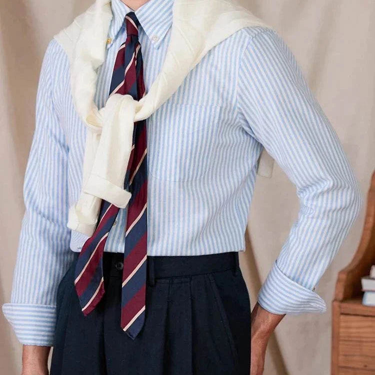 Oxford style casual cotton shirt with blue stripes, worn with a tie and sweater draped over shoulders.