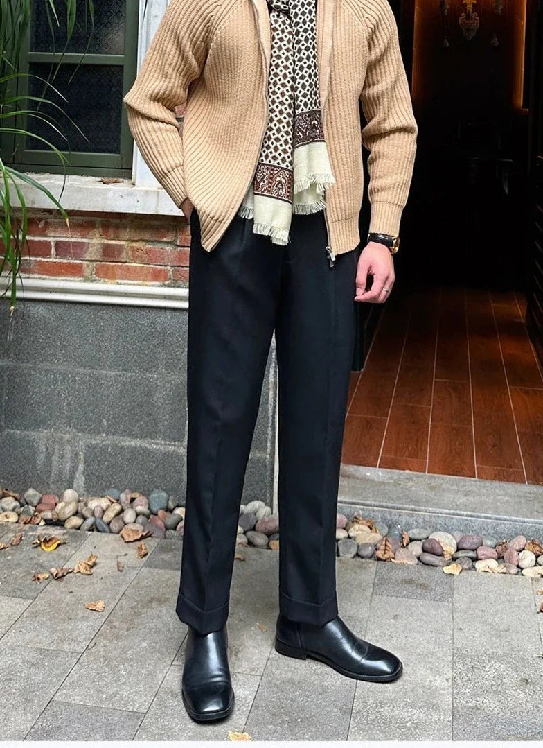 Man wearing retro cardigan with stylish black pants and boots, standing in front of brick wall.