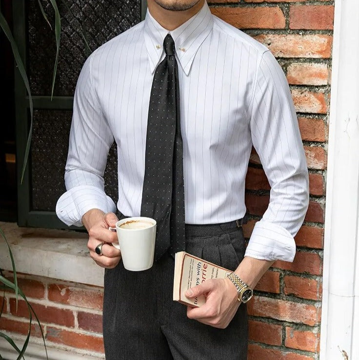 Man wearing Antonios Business Camisa with long sleeves and a tie, holding a coffee cup and book, against a brick wall background.