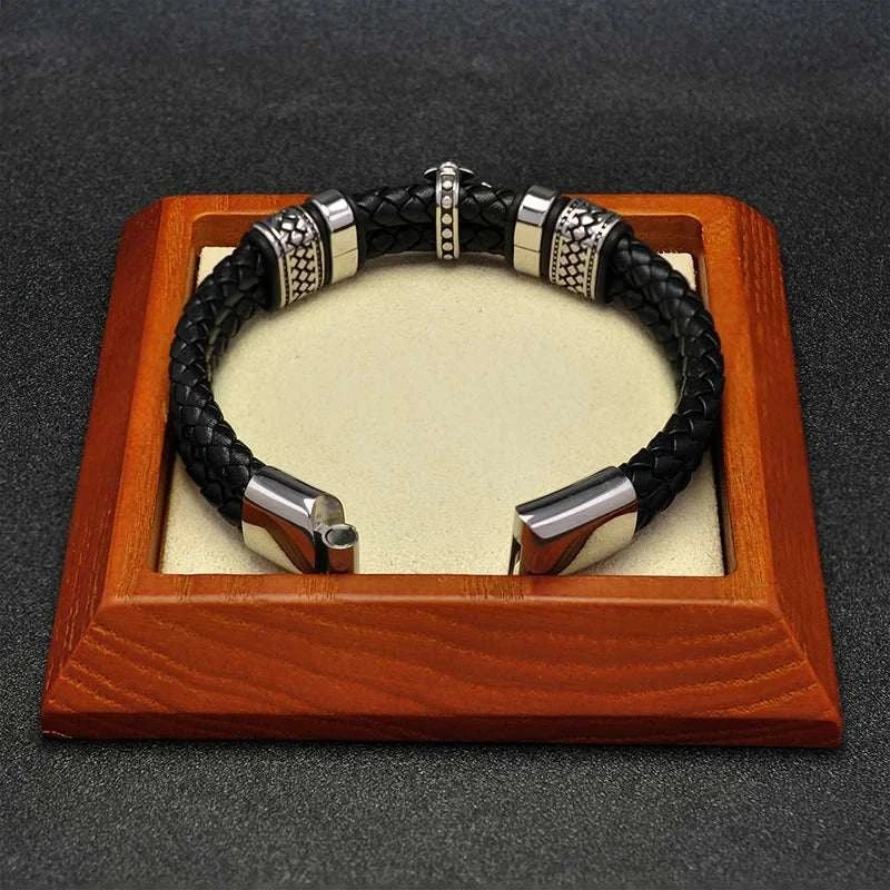Stainless steel cross magnet bracelet on a wooden stand with black braided band and metallic accents.