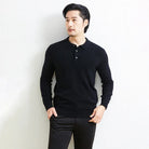 A man wearing a black long-sleeve polo knit sweater and black trousers, standing against a light textured wall.