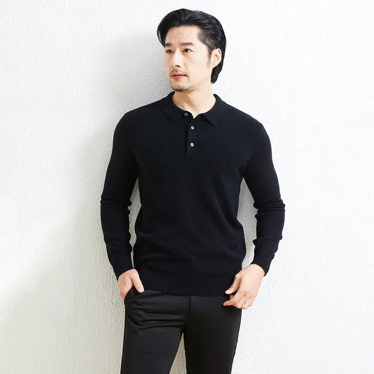 A man wearing a black long-sleeve polo knit sweater and black trousers, standing against a light textured wall.