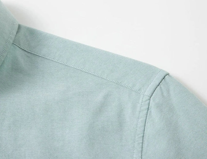 Close-up detail of a light sage green shirt showing the shoulder seam, collar, and fabric texture against a plain white background.