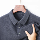A close-up of a dark gray heathered knit polo shirt hanging on a wooden hanger, with a hand holding the fabric to show its soft texture.