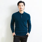 A man wearing a dark teal blue long-sleeve polo knit sweater and black trousers, standing against a light textured wall.