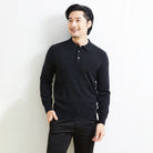 A man wearing a dark charcoal knit polo shirt and black pants, standing against a light textured wall.