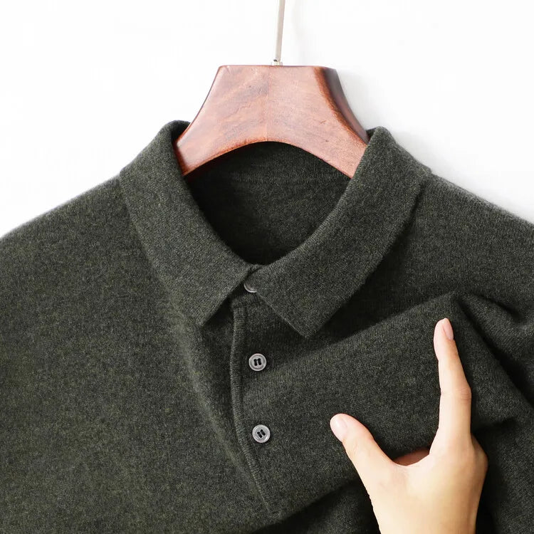 A close-up of a dark olive green knit polo shirt hanging on a wooden hanger against a white background, with a hand holding the fabric to show its texture.