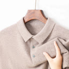 A close-up of a light taupe knit polo shirt hanging on a wooden hanger against a white background, with a hand holding the fabric to show its soft texture.