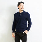 A man wearing a dark navy blue long-sleeve polo knit sweater and black trousers, standing against a light textured wall.